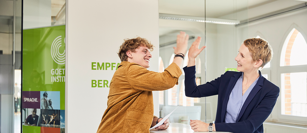 Two people smiling and high-fiving in an office setting, with a Goethe-Institut banner in the background.