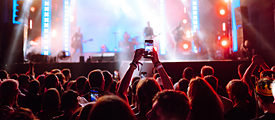 Blick aus dem Publikum auf eine hellerleuchtete Bühne. Eine Zuschauerin hält ein Mobiltelefon in die Höhe und filmt das Konzert.