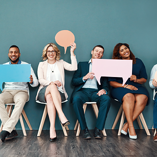 Des hommes et des femmes en tenue d'affaires, assis en ligne les uns à côté des autres, tiennent de grandes bulles colorées devant eux ou en l'air. Ils s'amusent et rient face à la caméra. Espace pour le texte rédactionnel.
