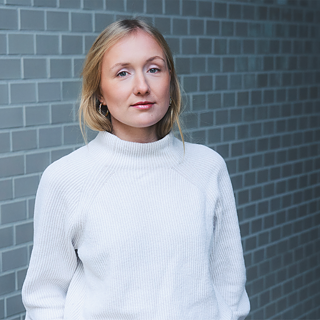 Foto af forfatteren Ruth-Maria Thomas, der smiler let foran en grå væg. 