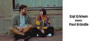 Ein Mann und eine Frau sitzen im Schneidersitz vor einer Hauswand und schauen einander an. Rechts neben dem Bild steht in schwarzer Schrift auf weißem Hintergrund "Enji Erkhem meets Paul Brändle".