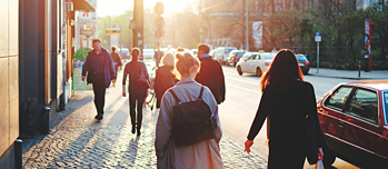People walking along a city sidewalk in the warm light of sunset, with cars passing by on the street.