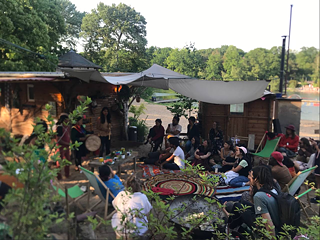 Das Bild zeigt eine Art Festival im Freien. Menschen sitzen auf Liegestühlen und am Boden und lauschen einer Musikgruppe. 