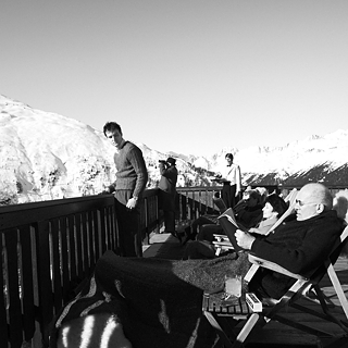 Mehrere Personen entspannen auf einer Sonnenterrasse in den Bergen, umgeben von Schnee und klarem Himmel.