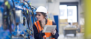 A person wearing a hard hat and safety vest inspects industrial machinery while holding a tablet.
