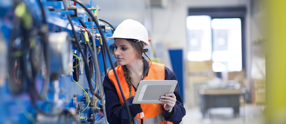 A person wearing a hard hat and safety vest inspects industrial machinery while holding a tablet.