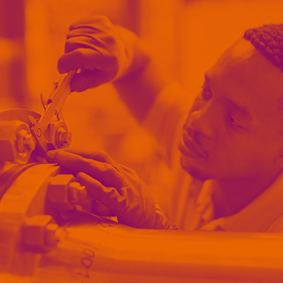 Männliche junge Person misst ein Teil an einer Maschine, trägt Arbeitshandschuhe, Bild in orangen und violetten Farbtönen