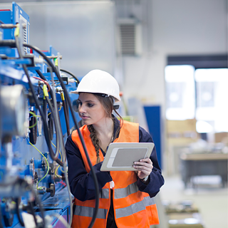 A person wearing a hard hat and safety vest inspects industrial machinery while holding a tablet.