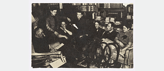 Familienbild der Familie Mann, Thomas Mann liest vor. Sie sitzen in einem Arbeitszimmer auf Stühlen und Sofas, im Hintergrund Bücherregale.  Von links nach rechts: Thomas Mann, Michael Mann, Katia Mann, Golo Mann, Erika Mann, Klaus Mann, Elisabeth Mann.
