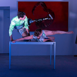 Melyname fone ant stalo jaunas vyras ir moteris šokio judesyje, viena vyro koja su protezu, už jų matyti tamsiai raudonos spalvos paveikslas