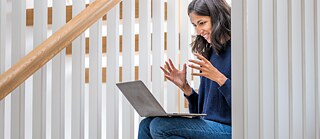 A woman sits in the stairwell. She communicates via the laptop. She is concentrated and smiles. She explains something and gestures with her hands.