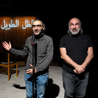 The image shows two actors on stage. One of them is speaking into a microphone and pointing to Arabic writing in the background.