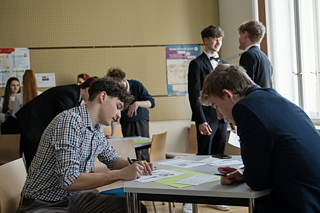 Mehrere Teilnehmer sitzen während des Sprachkurses an einem Tisch. Im Hintergrund unterhalten sich zwei weitere Teilnehmer.