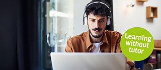A man sits facing a laptop, wearing headphones and looking focused at the screen while studying German through self-paced online learning.