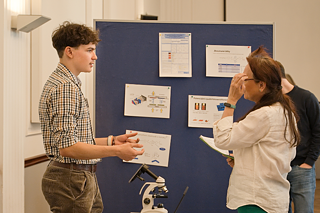 Ein Teilnehmer des Wettbewerbs erklärt vor einer Pinnwand, was er im Rahmen seines Projekts gemacht hat. Gesten unterstützen seine Aussagen. 