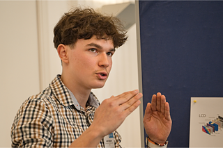 Ein Kandidat spricht zur Jury. Im Hintergrund ist eine Pinnwand zu sehen.