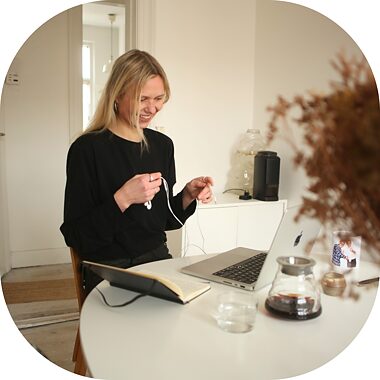 Eine junge Frau schaut lächelnd auf einen Computerbildschirm und hält ein paar Kopfhörer in der Hand.