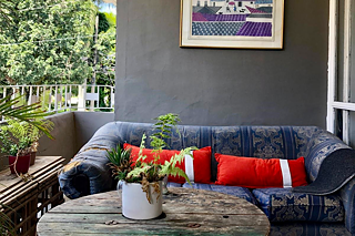 Auf einem Balkon stehen ein runder Holztisch und ein breites Sofa mit roten Kissen.