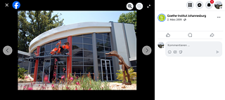 Das Bild zeigt den Eingangsbereich des Goethe-Instituts mit seinen großen Glasfenstern und einem geschwungenen Dach. Auf dem Vordach über der Eingangstür ist eine auffällige Skulptur die aus orange-rot-schwarzen Plastikstühlen gebaut wurde. Rechts im Bild, nahe einiger Pflanzen, steht eine weitere Skulptur – ein Dinosaurier aus Holz. Die Szene ist von Grünflächen umgeben und wirkt kreativ und einladend. Am rechten Bildrand befindet sich die Facbook-Kommentarspalte mit dem Text: „Goethe-Institut Johannesburg – 2. März 2009“