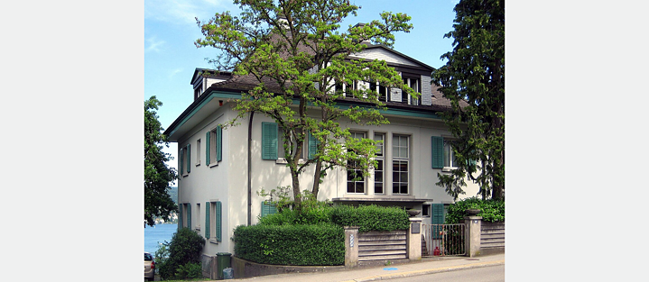 Ein großes zweistöckiges Haus mit heller Fassade und grünen Fensterläden. Das Haus hat ein schräges Dach mit mehreren Dachfenstern. Vor dem Haus steht ein Baum, der teilweise die Ansicht der Fassade verdeckt. Das Grundstück ist von Grünflächen umgeben, einschließlich Büschen und Bäumen, und es gibt einen Zaun mit einem Eingangstor vor dem Haus. Eine Straße verläuft vor dem Grundstück. In diesem Haus In Kilchberg am Zürichsee hat die Familie Mann von 1954 bis 1992 gelebt. Die Aufnahme ist von 2010.