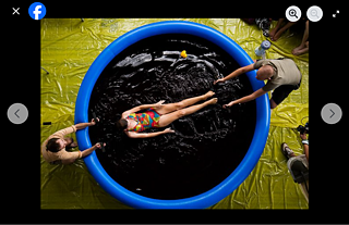 Das Bild zeigt eine Person in einem bunten Badeanzug, die auf dem Rücken in einem großen, runden, blauen aufblasbaren Pool treibt. Die Flüssigkeit im Pool scheint dunkel gefärbt. Zwei weitere Personen stehen außerhalb des Pools und halten jeweils eine Hand der im Wasser liegenden Person.  Der Boden rund um den Pool ist mit einer gelben Plane abgedeckt. Rechts im Bild befindet sich  die Fabeook-Kommentarspalte mit folgenden Informationen:  „Goethe-Institut Johannesburg – 2. März 2009 copyright Christo Doherty check out all the pics at http://www.flickr.com/search/...“