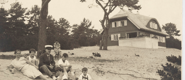 Ferienhaus der Familie Mann in Nidden, Litauen. Davor im Sand sitzend von links: Katia Mann, Elisabeth Mann, Thomas Mann, Michael Mann, unbekannter Junge, Alexander Ferber (Sohn des Mädchens Maria Ferber), auf der Bank im Hintergrund Monika Mann.
