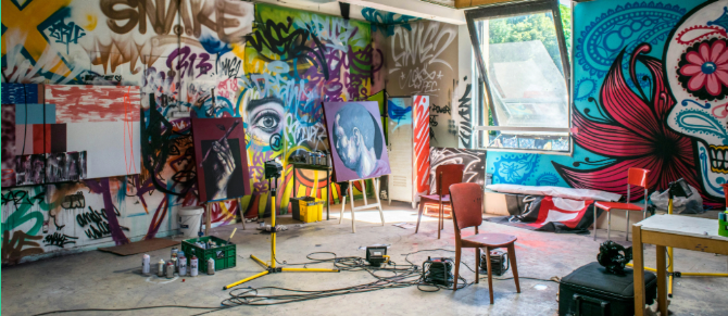 An art studio with graffiti-covered walls and an industrial interior. Several easels display paintings, including a large eye and a black-and-white portrait of two people. The space is filled with scattered art supplies, paint cans, and cables. Exposed pipes run along the ceiling, and natural light enters through an open window. Tables and chairs are arranged with more materials