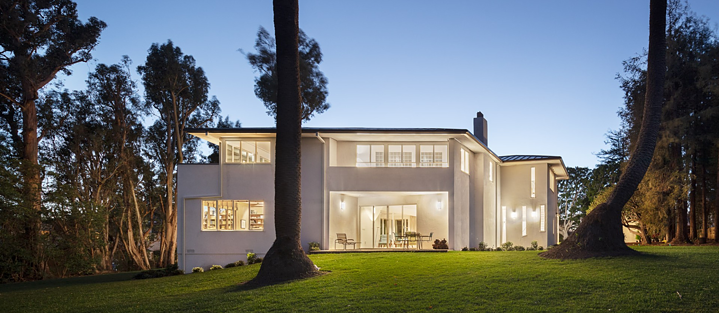 Das Thomas-Mann-House in Los Angeles, ein modernes zweistöckiges Gebäude mit weißer Fassade und großen Fenstern, umgeben von hohen Bäumen und einem gepflegten Rasen. Die Außenbeleuchtung und der Himmel bei Dämmerung schaffen eine ruhige Atmosphäre.