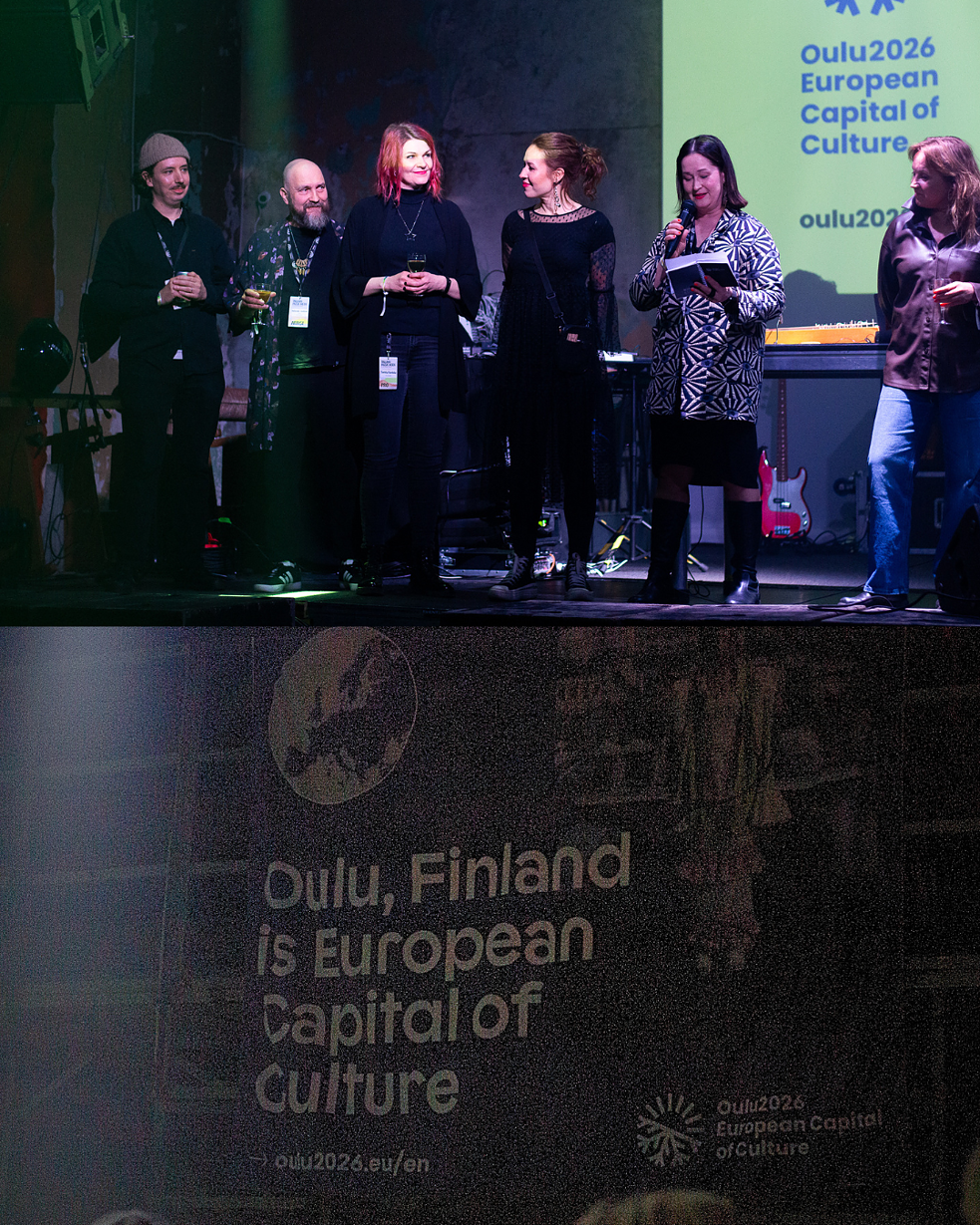 A group of people stands on a stage some holding microphones and others holding drinks.  Below the stage a banner reads: Oulu Finland is European Capital of Culture