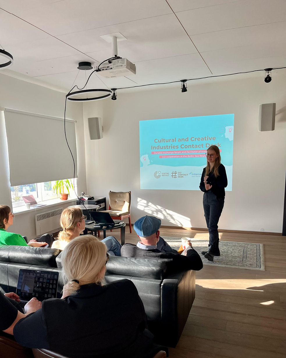 A group of people sits on couches and chairs in a well-lit room, watching a presentation. A presenter stands near a screen displaying the title: Cultural and Creative Industries Contact Day