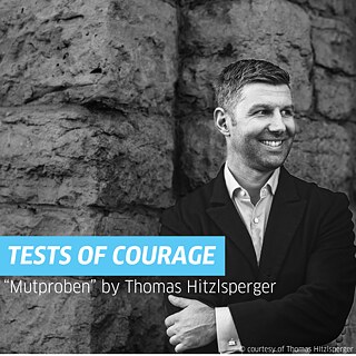 Portrait of Thomas Hitzlsperger wearing a black blazer and light blue striped shirt, standing in front of a brick building