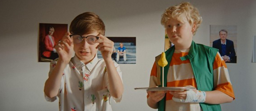 Two children standing indoors in front of a wall with three framed photographs. The child on the left wears a white shirt with colorful patterns and holds a pair of glasses near their face. The child on the right wears an orange and white striped shirt with a green vest, holding a tray with a yellow item.