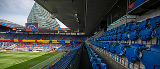 Blick aus den Zuschauerrängen in ein großes Fußballstadion mit blauen Sitzplätzen. Im Hintergrund sind bunte Banner und Fahnen sowie ein großes Gebäude außerhalb des Stadions zu sehen. Das Spielfeld ist grün und gepflegt, entlang der Seitenlinien sind Werbetafeln sichtbar.