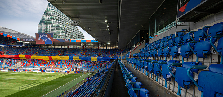 Blick aus den Zuschauerrängen in ein großes Fußballstadion mit blauen Sitzplätzen. Im Hintergrund sind bunte Banner und Fahnen sowie ein großes Gebäude außerhalb des Stadions zu sehen. Das Spielfeld ist grün und gepflegt, entlang der Seitenlinien sind Werbetafeln sichtbar.