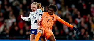 Zwei Fußballspielerinnen kämpfen während eines Spiels um den Ball. Die Spielerin links trägt ein weißes Trikot mit blauer Hose und der sichtbaren Nummer 12. Die Spielerin rechts trägt ein orangefarbenes Trikot mit passender Hose und der Nummer 7. Beide Gesichter sind unscharf dargestellt. Im Hintergrund ist eine Zuschauermenge im Stadion zu sehen.