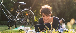 Eine junge Frau liegt mit ihrem Tablet neben ihrem Fahrrad auf einer grünen Wiese
