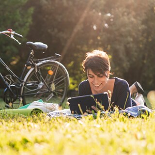 Eine junge Frau liegt mit ihrem Tablet neben ihrem Fahrrad auf einer grünen Wiese