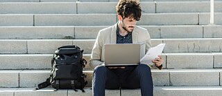 Junger Mann lernt am Laptop auf einer Treppe im Freien mit Rucksack nebenan