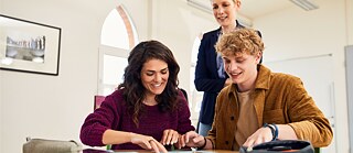 Eine junge Frau und ein junger Mann sitzen an einem Schreibtisch in einem Klassenraum mit einer Lehrkraft im Hintergrund