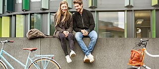 Eine junge Frau und ein junger Mann sitzen lächelnd zwischen zwei geparkten Fahrrädern