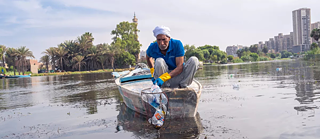 رجل مسن يجلس في مركب على نهر النيل ويجمع البلاستيك من المياه بشبكة يدوية.