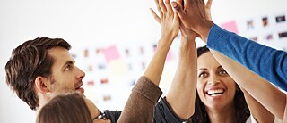 Several colleagues at a meeting in the office. They high-five and are smiling.