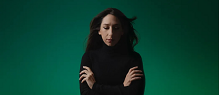 Eine Frau steht vor einem grünen Hintergrund und schaut nach unten. Ihre Hände hält sie gekreuzt vor ihrer Brust. Ihre Haare sind verweht. Über ihr schwebt ein hellgrün leuchtendes Ufo.