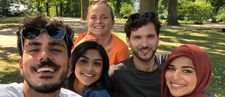 A group of five diverse friends are posing for a selfie in a park on a sunny day. They are smiling and looking at the camera. One woman is wearing a hijab. Trees and green space fill the background.