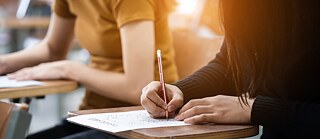 A student diligently writing on paper at a desk, with a notebook and pen in hand, deep in thought
