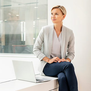 A woman is sitting in an office on a window sill in front of a modern glass façade, looking out of the window. There is a laptop in front of her.