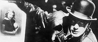 Foto en blanco y negro del clásico del cine alemán M de Fritz Lang: Un hombre con sombrero y abrigo de cuero sostiene la foto de una niña. Los rostros de ambas personas están difuminados.