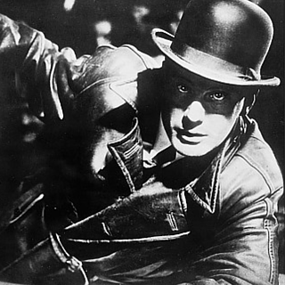 Foto en blanco y negro del clásico del cine alemán M de Fritz Lang: Un hombre con sombrero y abrigo de cuero sostiene la foto de una niña. Los rostros de ambas personas están difuminados.