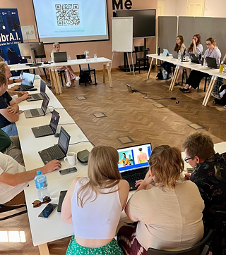 Das Bild zeigt eine Gruppe von Menschen, die an einer Schulung in einem gut beleuchteten Raum mit Holzboden teilnehmen. Sie sitzen um eine große U-förmige Anordnung weißer Tische herum, wobei jede Person an einem Laptop arbeitet. Im Vordergrund betrachten drei Teilnehmer aufmerksam einen Laptop-Bildschirm, auf dem KI-generierte Bilder von Menschen angezeigt werden. Auf der linken Seite des Bildes sitzt eine Frau vorne im Raum an einem Tisch und leitet die Sitzung. Hinter ihr befindet sich eine Projektionsfläche, auf der ein QR-Code und ein Link zu sehen sind. Auf der linken Seite des Raums ist ein Banner des LIBRA.I.-Projekts zu sehen. An der Rückwand stehen ein großer Monitor, ein Flipchart und einige Stühle. Die Atmosphäre wirkt konzentriert und kooperativ.