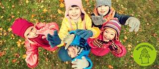 Eine Gruppe von Kindern hebt im Park lächelnd die Hände – sie tragen Winterkleidung.